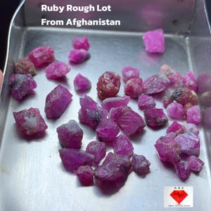 Puede incluir: Un conjunto de gemas de rubí en bruto, en varios tonos de rosa y rojo, presentadas en una bandeja metálica. El texto indica que el lote pesa 33,45 ct y es de Afganistán.