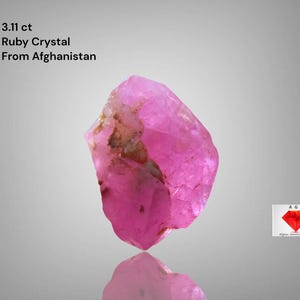 May include: A vivid pink ruby crystal from Afghanistan, weighing 3.11 ct. The rough gemstone has a natural, irregular shape with a reflective surface. The image includes text indicating the crystal's origin and weight.