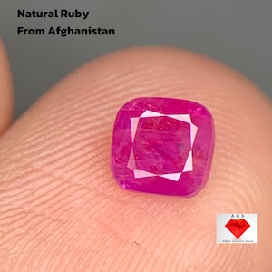 Può includere: Un rubino naturale taglio a cuscino dall'Afghanistan, 0,610 ct. La gemma è di un vivace colore rosso rosato, di forma quadrata e con un motivo interno bianco. Il testo "0.610 ct Natural Ruby From Afghanistan" è presente.