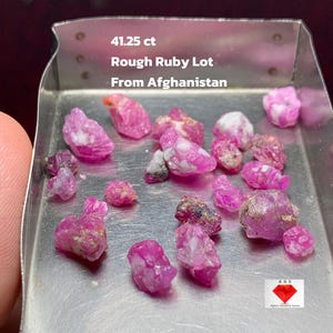 Puede incluir: Un lote de gemas de rubí en bruto, con un peso total de 41,25 ct, exhibido en una bandeja metálica. Los rubíes son de varios tonos de rosa y rojo, con algunas inclusiones blancas. El texto dice: "Rough Ruby Lot From Afghanistan."