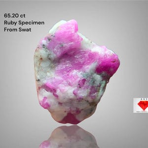 May include: A 65.20 ct ruby specimen from Swat, Pakistan. The gemstone displays a vibrant pink colour with white and grey inclusions. The rough ruby is irregularly shaped and reflects light, showcasing its natural beauty.