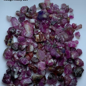 Op de afbeelding: Een verzameling ruwe robijn edelstenen, met een gewicht van 165,10 ct. De stenen vertonen een reeks kleuren, waaronder diep rood, paars en roze. De tekst "Rough Ruby Lot" is zichtbaar.