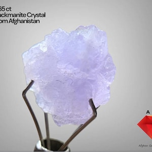 Puede incluir: Un cristal de hackmanita de color púrpura claro de Afganistán, sostenido por un soporte metálico. El texto dice "5.65 ct Hackmanite Crystal From Afghanistan". El cristal tiene una superficie rugosa y texturizada y es translúcido. El logotipo de "Afghan Gemstone Source" está en la parte inferior derecha.