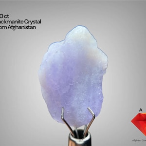 Könnte beinhalten: Ein 9,10 ct Hackmanit-Kristall aus Afghanistan mit einer blassvioletten, durchscheinenden und unregelmäßigen Kristallstruktur, gehalten von einem metallenen Krallenständer. Der Text 9.10 ct Hackmanite Crystal From Afghanistan ist sichtbar.