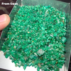 Puede incluir: Una bandeja llena de un lote de esmeraldas en bruto. Las esmeraldas son de color verde vibrante y parecen sin cortar. El texto en la imagen dice "323.65 ct Emerald Rough Lot From Swat."