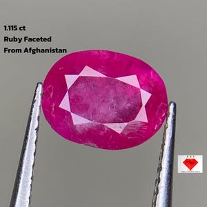 May include: A faceted ruby gemstone from Afghanistan, held by tweezers. The oval-shaped gem is a vibrant pinkish-red with a star-like pattern in the center. Text on the image reads: "1.115 ct Ruby Faceted From Afghanistan."