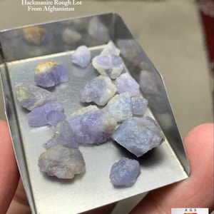 May include: A collection of rough-cut hackmanite gemstones from Afghanistan. The stones are a pale lavender color and are displayed in a small, silver tray. Text on the image reads "42.85 ct Hackmanite Rough Lot From Afghanistan."