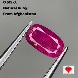 Puede incluir: Un rubí natural de Afganistán, de 0,615 ct, es sujetado por unas pinzas sobre un fondo gris. La gema es de un rojo intenso con un corte rectangular y un patrón geométrico blanco.