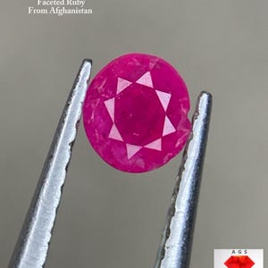 May include: A faceted, round, deep pink ruby gemstone from Afghanistan is held by tweezers. The gem has a star-like pattern of facets. Text at the top reads "0.260 ct Faceted Ruby From Afghanistan". A logo is in the bottom right corner.