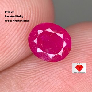 Puede incluir: Un rubí facetado de 1,110 ct, de Afganistán. La gema es de color rojo vibrante con un patrón de facetas en forma de estrella. La imagen también incluye el logotipo de AGS.