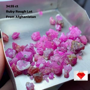 May include: A collection of rough-cut ruby gemstones, predominantly pink and red, displayed in a small metal tray. The text "34.55 ct Ruby Rough Lot From Afghanistan" is visible. The image also includes a logo for "AGS Afghan Gemstone Source."