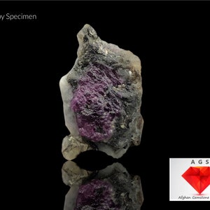 Peut inclure: Un spécimen brut de rubis gris foncé et violet avec une matrice blanche. Le spécimen est sur un fond noir et réfléchit la lumière. Le texte "AGS Afghan Gemstone Source" est dans le coin inférieur droit de l'image.