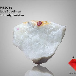 Peut inclure: Spécimen de rubis blanc et beige d'Afghanistan, pesant 441,20 ct. La pierre brute présente un petit cristal de rubis rouge vif incrusté au centre. L'image comprend le texte "Ruby Specimen From Afghanistan."