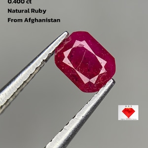 Puede incluir: Un rubí natural de corte rectangular, sujeto con pinzas. La piedra de color rojo intenso, con un peso de 0,400 ct, es de Afganistán. El texto en la imagen dice "Natural Ruby From Afghanistan".