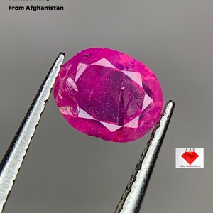 Peut inclure: Un rubis facetté de forme ovale, tenu par une pince. Le rubis est d'un rouge-rosé profond. Le texte de l'image indique : "0,75 ct Rubis Facetté D'Afghanistan". Un logo avec le texte "AGS Afghan Gemstone Source" est dans le coin inférieur droit.
