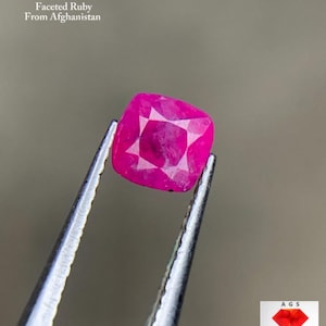 Könnte beinhalten: Ein facettierter Rubin im Kissenschliff, gehalten von einer Pinzette. Der Rubin hat eine leuchtende rosa-rote Farbe mit sichtbaren Facetten. Der Text im Bild lautet "0,56 ct Faceted Ruby From Afghanistan". Ein Logo befindet sich unten rechts.