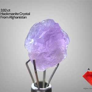 May include: A light purple Hackmanite crystal from Afghanistan, weighing 3.92 ct, is held by a metal stand. The crystal has a rough, textured surface. Text on the image reads "3.92 ct Hackmanite Crystal From Afghanistan".