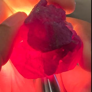 Puede incluir: Una piedra preciosa de rubí en bruto de forma irregular con un tono rojo intenso y vibrante. La piedra es translúcida, lo que permite que una fuente de luz brillante desde atrás ilumine su estructura interna, revelando texturas y formaciones cristalinas.