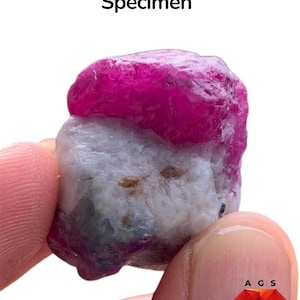 May include: A 110 ct ruby specimen featuring a vibrant, deep pinkish-red crystalline section atop a textured, opaque white and grey mineral matrix with small brown inclusions. The text 110 ct Ruby Specimen is visible above the stone.