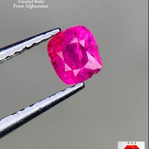 Pode incluir: Uma gema de rubi rosa vibrante e facetada, segurada por uma pinça. O texto diz "0,25 ct Faceted Ruby From Afghanistan". A gema tem formato quadrado com um padrão de estrela. Inclui o logotipo da Afghan Gemstone Source.