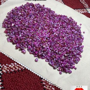 Pode incluir: Um monte de pedras preciosas de rubi em bruto, com um peso total de 2580 ct, do Afeganistão. Os rubis são de uma cor vermelho-rosada profunda e parecem não polidos. O texto "Ruby Rough Lot" é visível.