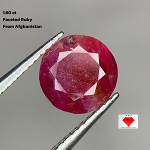 Puede incluir: Un rubí facetado, sostenido por unas pinzas, con el texto "1.60 ct Faceted Ruby From Afghanistan". El rubí es de color rojo intenso con un patrón de facetas en forma de estrella en el centro. La gema es redonda.