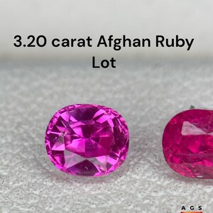 Pode incluir: Close-up de dois rubis afegãos rosa-avermelhados, lapidação oval e facetados. O texto diz "3.20 carat Afghan Ruby Lot". As pedras preciosas são apresentadas sobre uma superfície texturizada cinza claro. O logotipo "Afghan Gemstone Source" é visível no canto inferior direito.