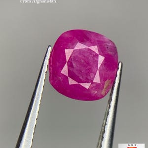 Puede incluir: Un rubí facetado de Afganistán, sujeto con pinzas. El rubí es de color rosa vibrante con un patrón en forma de estrella en el centro. El texto dice "2.07 ct Faceted Ruby From Afghanistan."