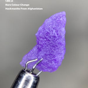 Puede incluir: Una vibrante gema de hackmanita púrpura de Afganistán, sujeta por una herramienta de metal. El texto de la imagen dice "1.80 ct Rare Colour Change Hackmanite From Afghanistan". La gema tiene una superficie rugosa y texturizada.