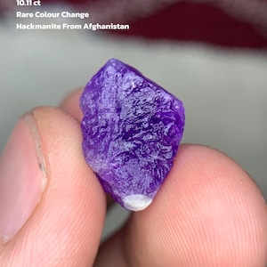 Puede incluir: Una vibrante gema de hackmanita púrpura de Afganistán. La piedra en bruto muestra un raro cambio de color y pesa 10,11 ct. El texto de la imagen dice "Cambio de color raro" y "Hackmanita de Afganistán."