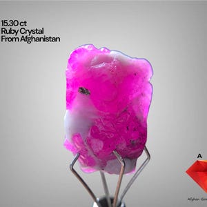 Puede incluir: Un vibrante cristal de rubí rosa de Afganistán, con un peso de 15,30 ct, se exhibe sobre un soporte plateado. La gema presenta una mezcla de tonos rosas y blancos, con el texto "15.30 ct Ruby Crystal From Afghanistan" visible.