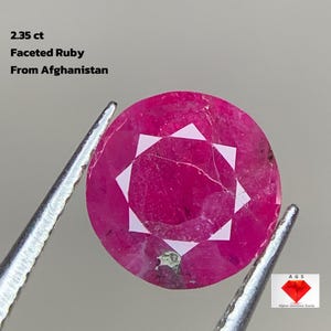 Puede incluir: Un rubí facetado, sostenido por pinzas, con un tono rosa vibrante y un patrón de estrella. El texto dice "2.35 ct Rubí Facetado De Afganistán". Un logotipo de "Afghan Gemstone Source" está en la esquina.