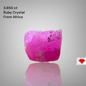Puede incluir: Cristal de rubí rosa vibrante de África, con un peso de 3,850 ct. La gema en bruto tiene una superficie reflectante y se muestra sobre un fondo gris claro. La imagen incluye texto que indica el origen y el peso del cristal.