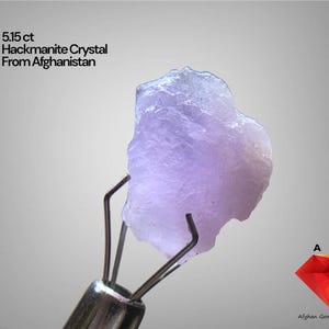 Puede incluir: Un cristal de hackmanita de 5,15 ct de Afganistán, sujeto con pinzas metálicas. El cristal es de color púrpura pálido con una forma rugosa e irregular. El texto de la imagen dice "5,15 ct Hackmanite Crystal From Afghanistan".