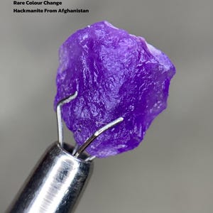 Puede incluir: Una vibrante gema de Hackmanita púrpura de Afganistán, sujeta por una herramienta plateada. La imagen incluye el texto "3.01 ct Rare Colour Change Hackmanite From Afghanistan."