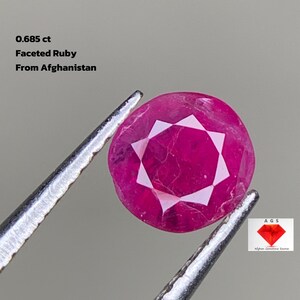 Puede incluir: Un rubí facetado, sostenido por unas pinzas, con un peso de 0,685 ct. La gema es de color rojo vibrante con un patrón de estrella. El texto en la imagen dice "Faceted Ruby From Afghanistan."