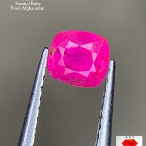 Puede incluir: Un rubí facetado de color rosa brillante, sujeto con pinzas. La gema es de forma cuadrada. El texto en la parte superior dice "0.40 ct Faceted Ruby From Afghanistan". La imagen incluye un logotipo en la esquina inferior derecha.