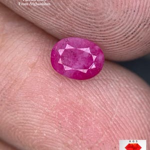 May include: A faceted, oval ruby gemstone, weighing 0.67 ct, from Afghanistan. The ruby is a vibrant pinkish-red colour with visible facets. The image also includes the text "0.67 ct Faceted Ruby From Afghanistan".