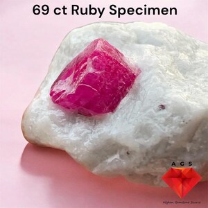 Puede incluir: Un espécimen de rubí de 69 ct con una estructura cristalina de color rojo rosado intenso incrustada en una matriz mineral blanca, rugosa y opaca. El texto 69 ct Ruby Specimen es visible arriba, con un logotipo A G S abajo a la derecha.