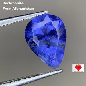 May include: A pear-shaped, deep blue hackmanite gemstone from Afghanistan, held by tweezers. The gem is 1.31 ct and has a faceted surface. The text "1.31 ct Hackmanite From Afghanistan" is visible. A logo for Afghan Gemstone Source is in the corner.