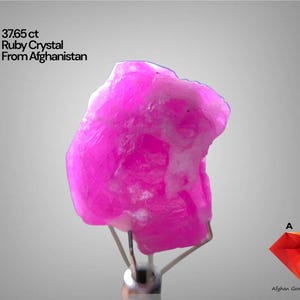 Puede incluir: Cristal de rubí rosa vibrante de Afganistán, con un peso de 37,65 ct. La gema está montada sobre un soporte plateado sobre un fondo gris claro. Incluye el texto "37.65 ct Ruby Crystal From Afghanistan".