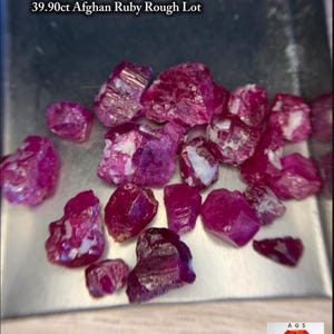 Puede incluir: Una colección de rubíes en bruto sin pulir con tonos rojo intenso y magenta, que presentan formas irregulares y texturas cristalinas. El texto 39.90ct Afghan Ruby Rough Lot es visible arriba y un logotipo AGS está en la esquina.