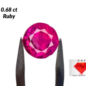 May include: A 0.68 carat ruby gemstone, a vibrant pink color, held in tweezers. The ruby is round and faceted. The image also includes a logo for Afghan Gemstone Source.
