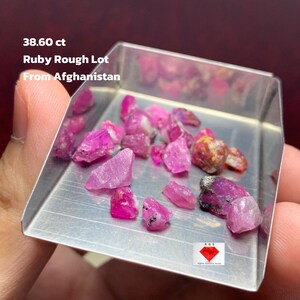 May include: A collection of raw, uncut ruby gemstones in a small, rectangular metal tray. The rubies are various shades of pink and red. Text on the image reads "38.60 ct Ruby Rough Lot From Afghanistan."