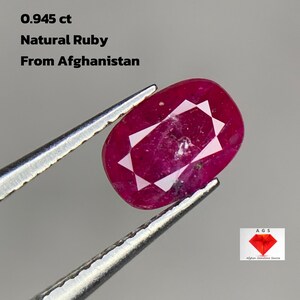 Może przedstawiać: Owalny, naturalny rubin z Afganistanu, trzymany pęsetą. Kamień szlachetny jest w głębokim odcieniu czerwieni z wzorem gwiazdy. Tekst: 0,945 ct, Natural Ruby, From Afghanistan. Logo w prawym dolnym rogu.