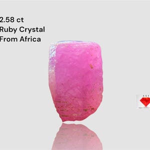 Puede incluir: Un vibrante cristal de rubí rosa de África, con un peso de 2,58 quilates. La gema en bruto tiene una superficie rugosa y texturizada y un aspecto natural y sin pulir. La imagen incluye texto que indica el origen y el peso del cristal.