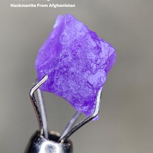 Puede incluir: Una vibrante gema de hackmanita púrpura de Afganistán, sujeta por una herramienta de metal. El texto dice "2.57 ct Rare Colour Change Hackmanite From Afghanistan". La gema tiene una superficie rugosa y texturizada.