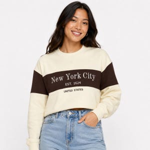 May include: A cream-colored long-sleeve sweatshirt featuring a wide horizontal dark brown stripe across the chest. The text New York City, EST. 1624, and UNITED STATES is printed in white on the dark brown stripe. The sweatshirt has a crew neckline.