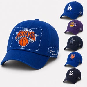 May include: A collection of six baseball caps in various colors, each featuring a sports team logo embroidered on the front and the text Your Name embroidered on the side. The caps include designs for the Knicks, Dodgers, Lakers, Cowboys, Mets, and Yankees.