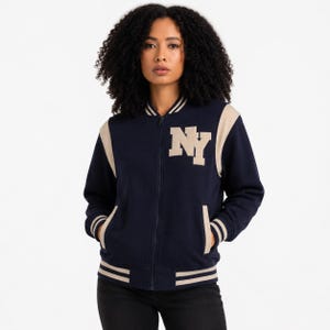 May include: A navy blue varsity-style bomber jacket featuring a full-length front zipper, beige sleeves, and beige striped ribbed trim at the collar, cuffs, and hem. The letters NY are appliqued in large beige block font on the upper left chest.
